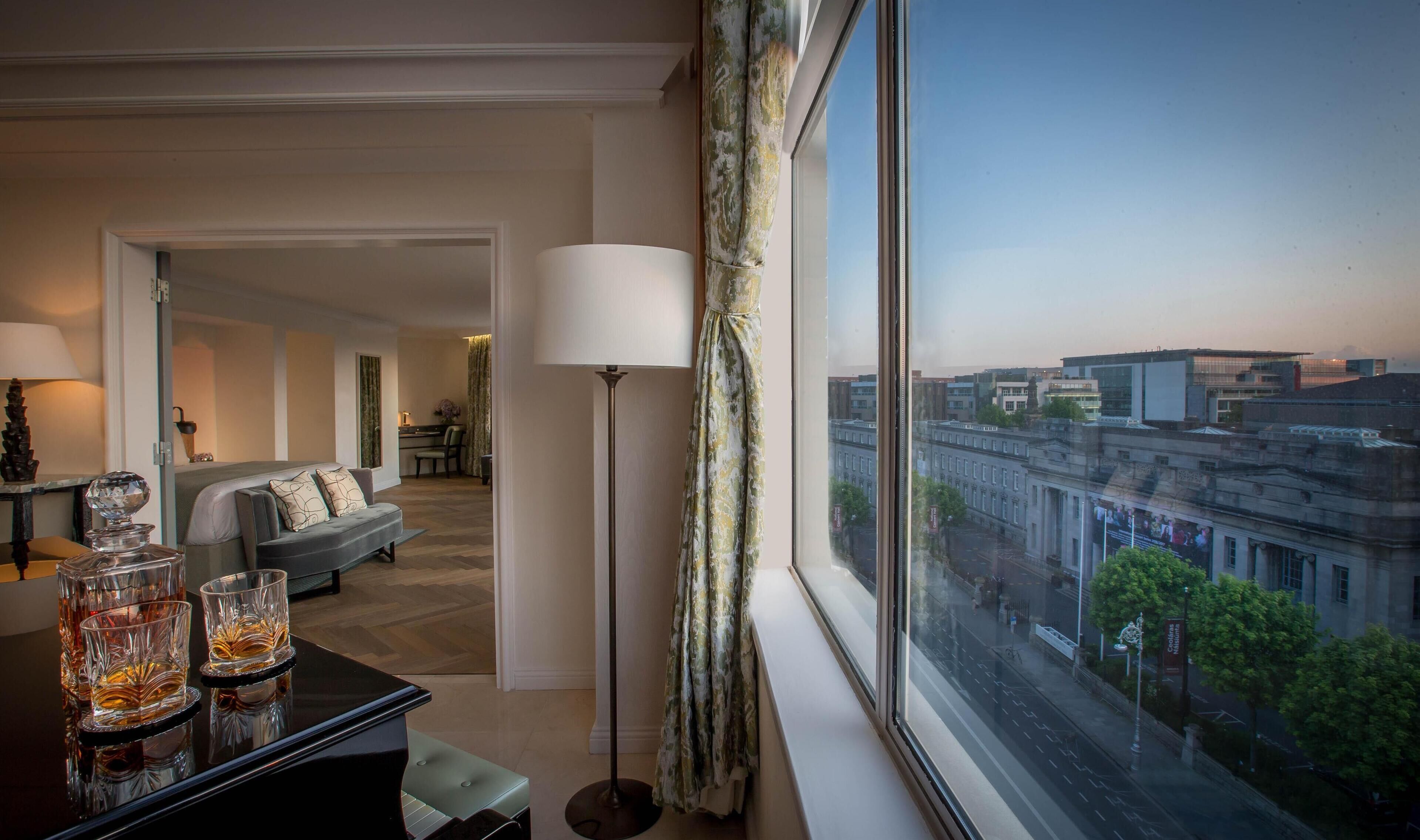 Une chambre d’hôtel avec vue sur la ville, un canapé, une table à manger avec des verres et une lampe.