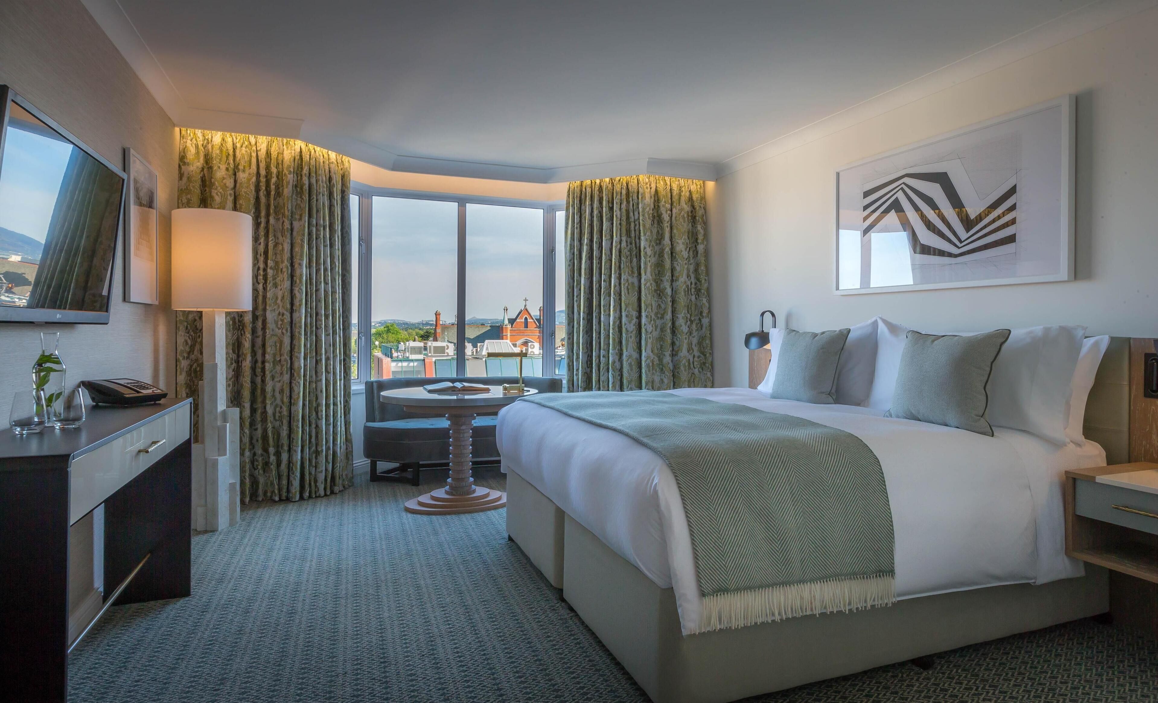 A hotel room with a bed, a desk, a chair, a small table, and a TV. The room has a view of a cityscape through large windows.