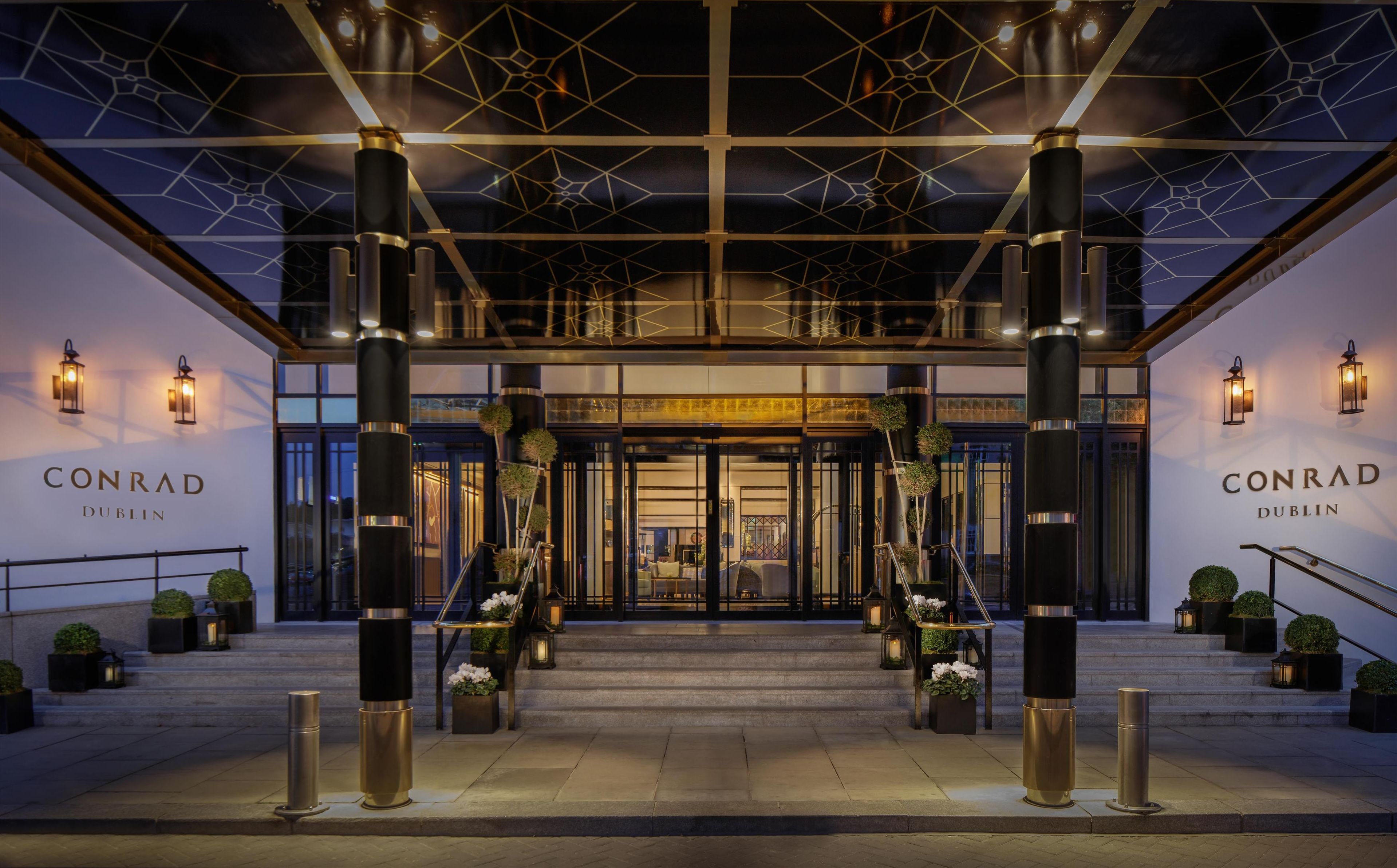De ingang van hotel Conrad Dublin met een modern ontwerp, met grote pilaren, een geometrisch plafond en een uitnodigende trap.