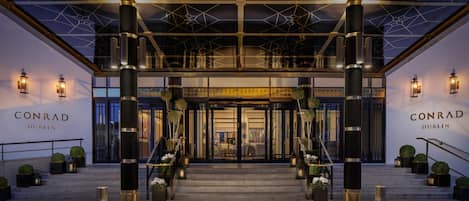 The entrance of Conrad Dublin hotel with a modern design, featuring large pillars, a geometric ceiling, and a welcoming staircase.