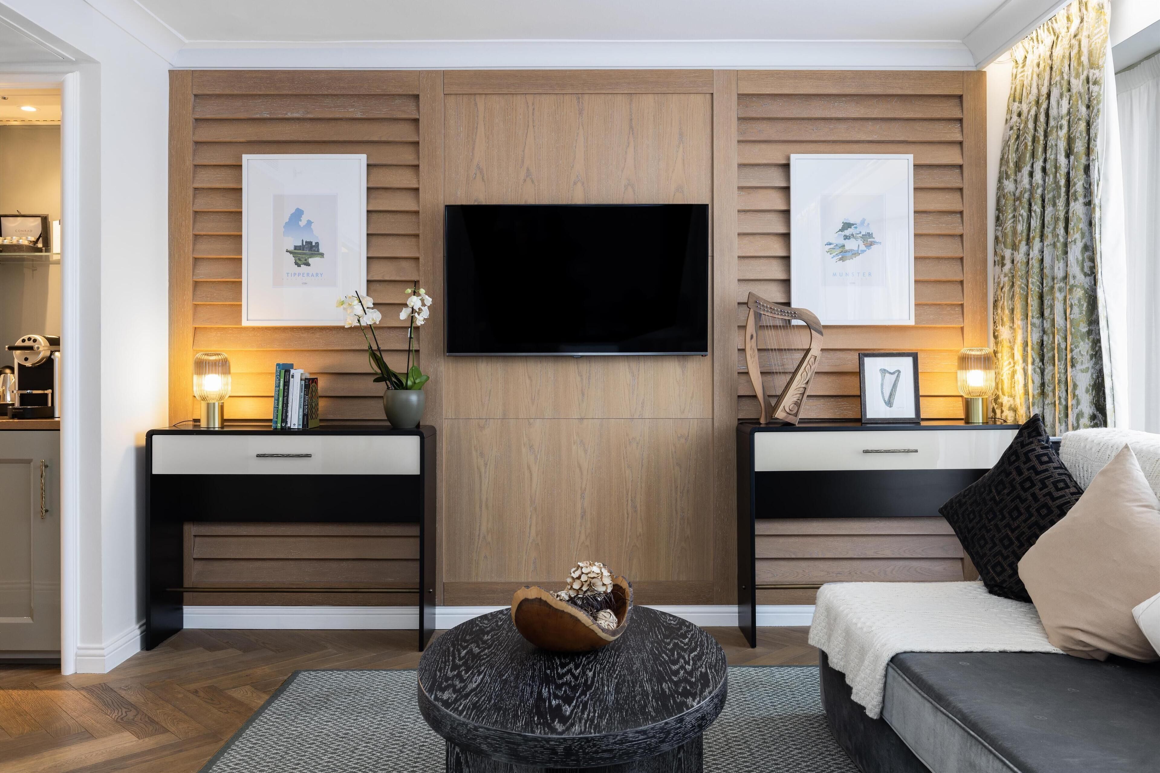 Un salon moderne avec un téléviseur à écran plat fixé à un mur en bois, deux tables d’appoint ornées d’objets décoratifs, une table basse ronde et un canapé avec des coussins.