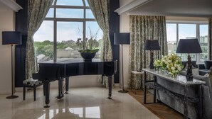 A grand piano in a spacious room with a large window, a table with a vase of flowers, and a lamp. - Conrad Dublin (Dublin)