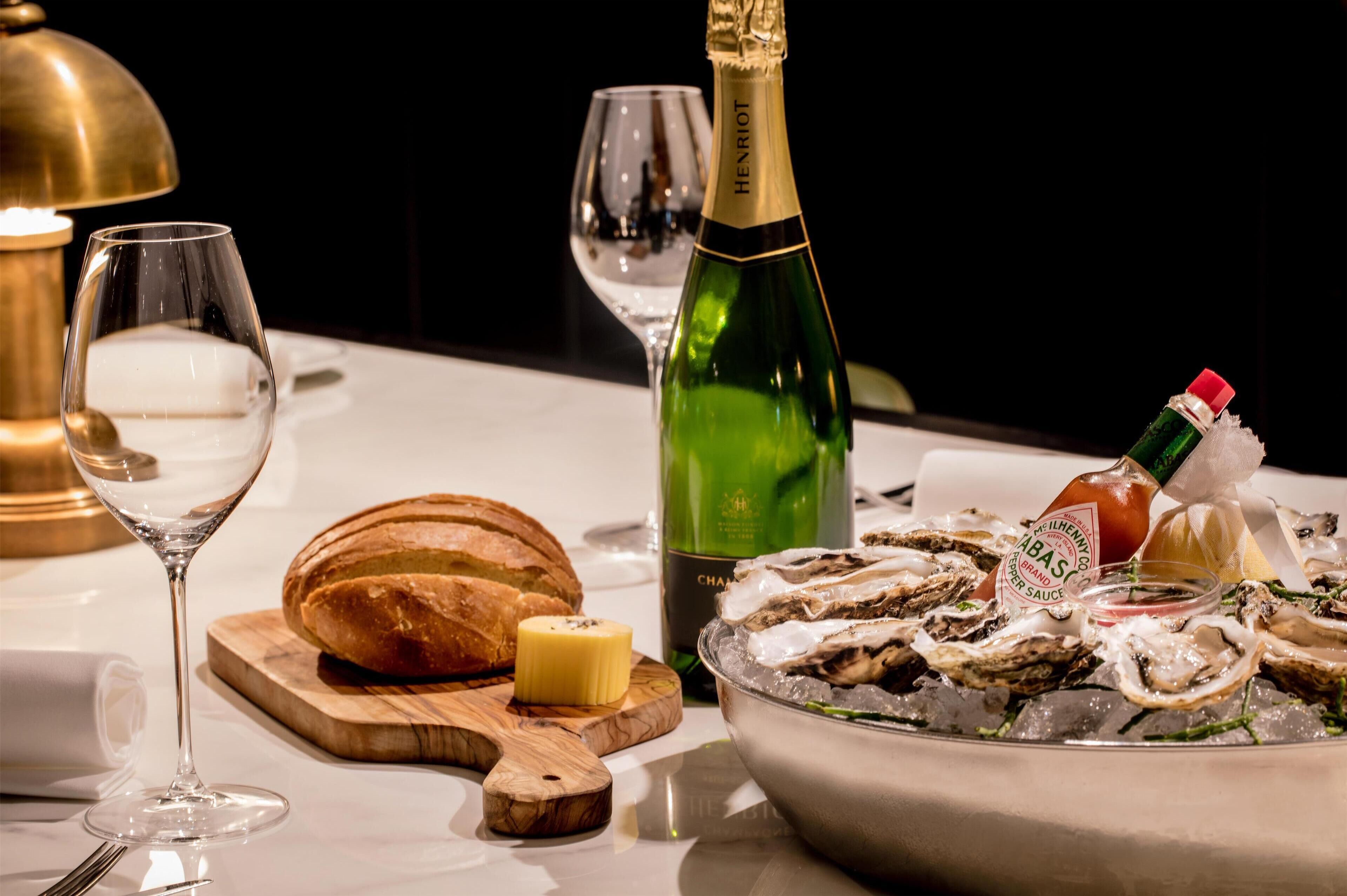 Una mesa con una botella de champán, una copa de vino blanco, una hogaza de pan, un recipiente pequeño de mantequilla y un bol de ostras sobre hielo.
