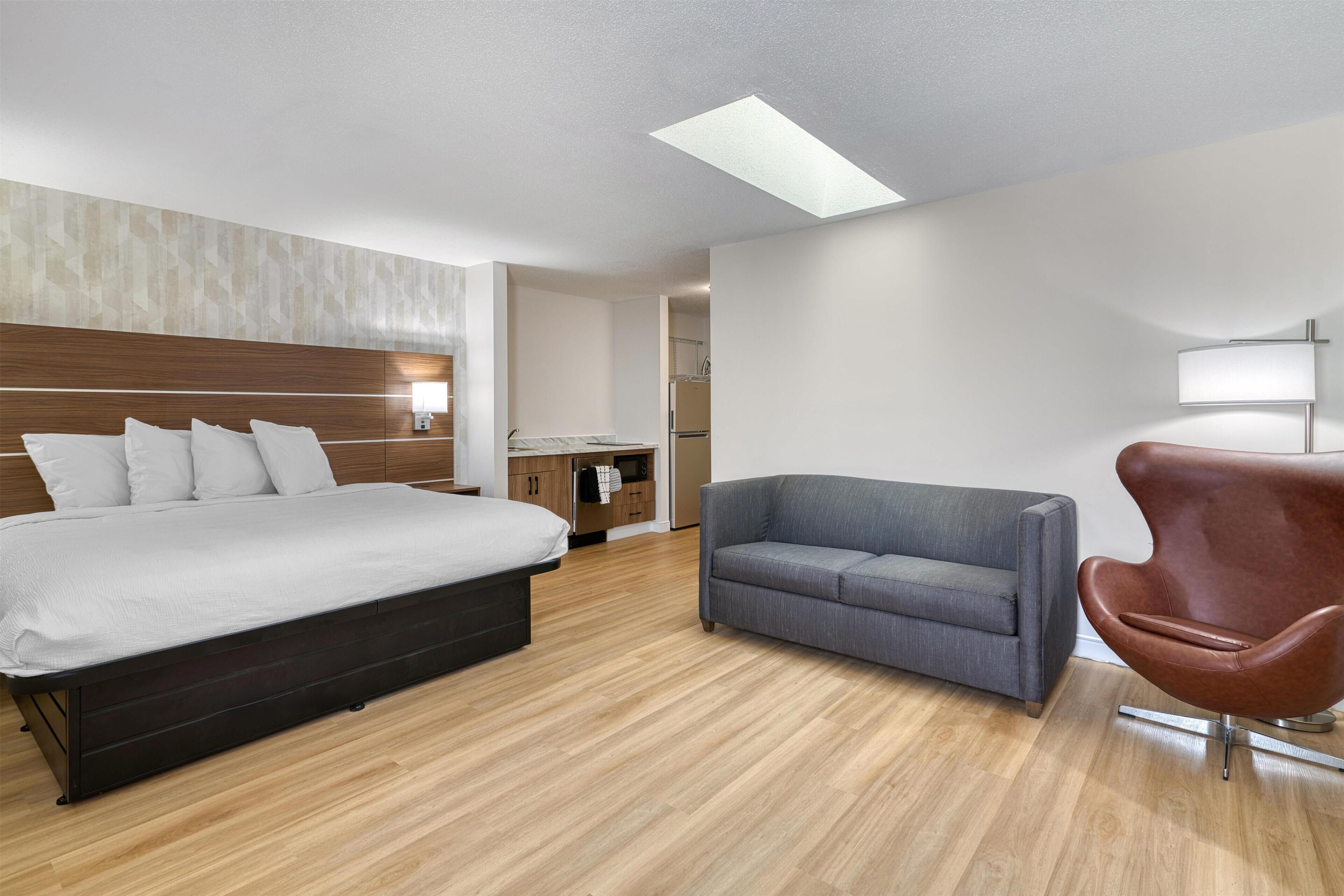 Quarto de hotel moderno com uma cama grande, um sofá cinzento, uma poltrona marrom e um claraboia.