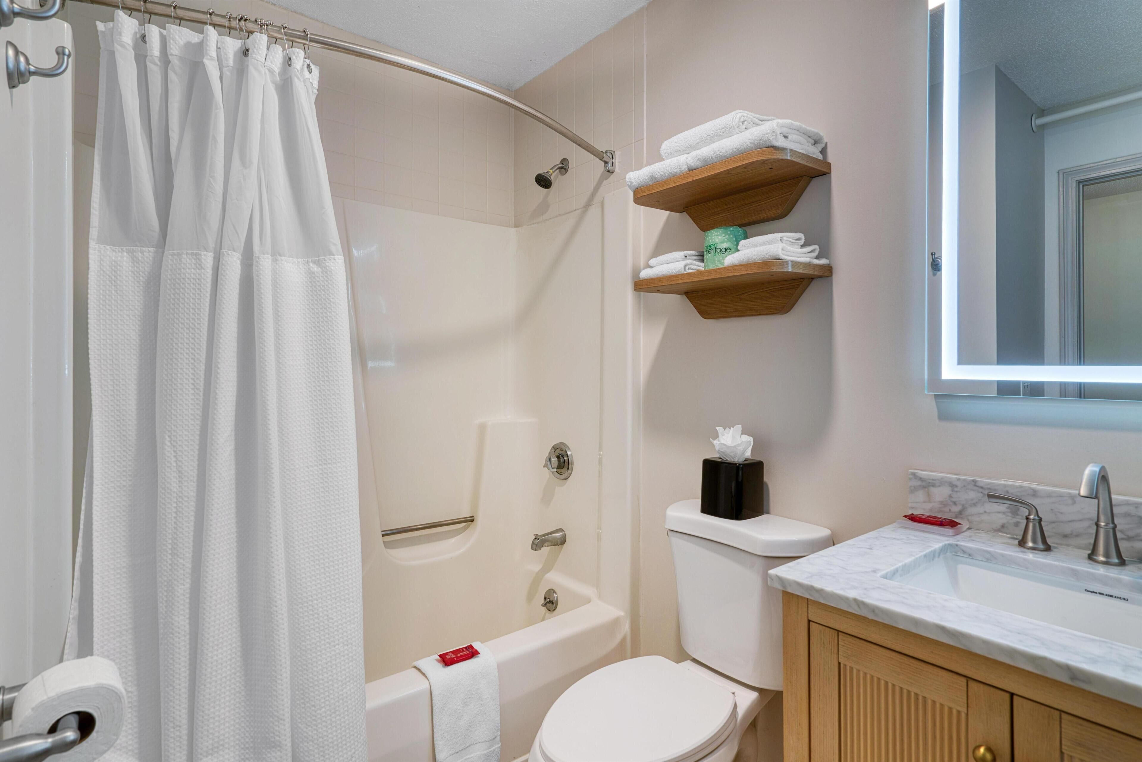 Ein Badezimmer mit Duschvorhang, Badewanne, Toilette und Waschbecken mit Marmorplatte. Holzregale mit Handtüchern.