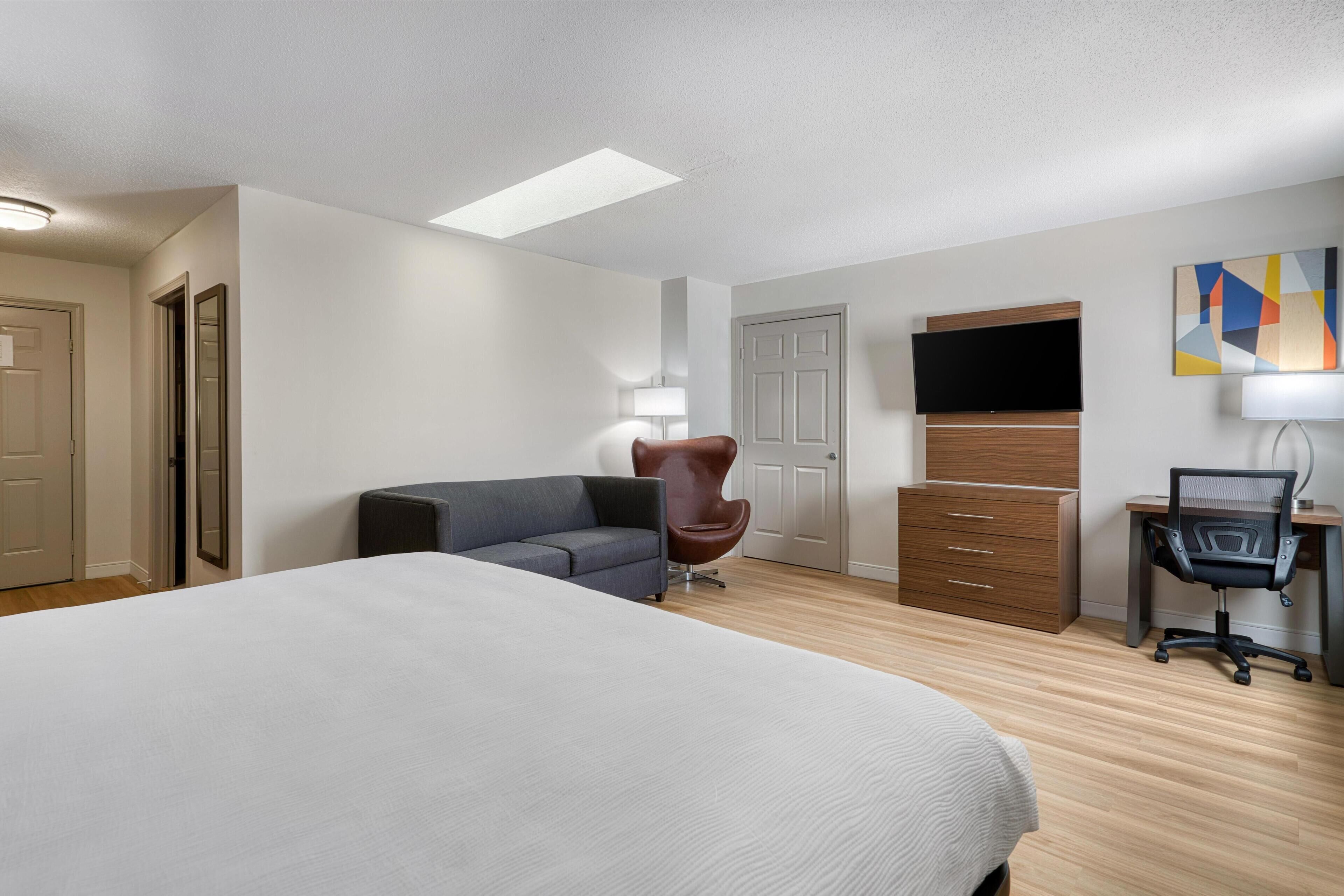 Habitación de hotel con una cama grande, un escritorio con una silla, un sillón de cuero marrón, un televisor montado en la pared y un cuadro sobre el escritorio.