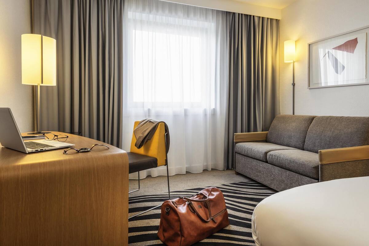 A hotel room with a desk, laptop, chair, sofa, and a large window with curtains.