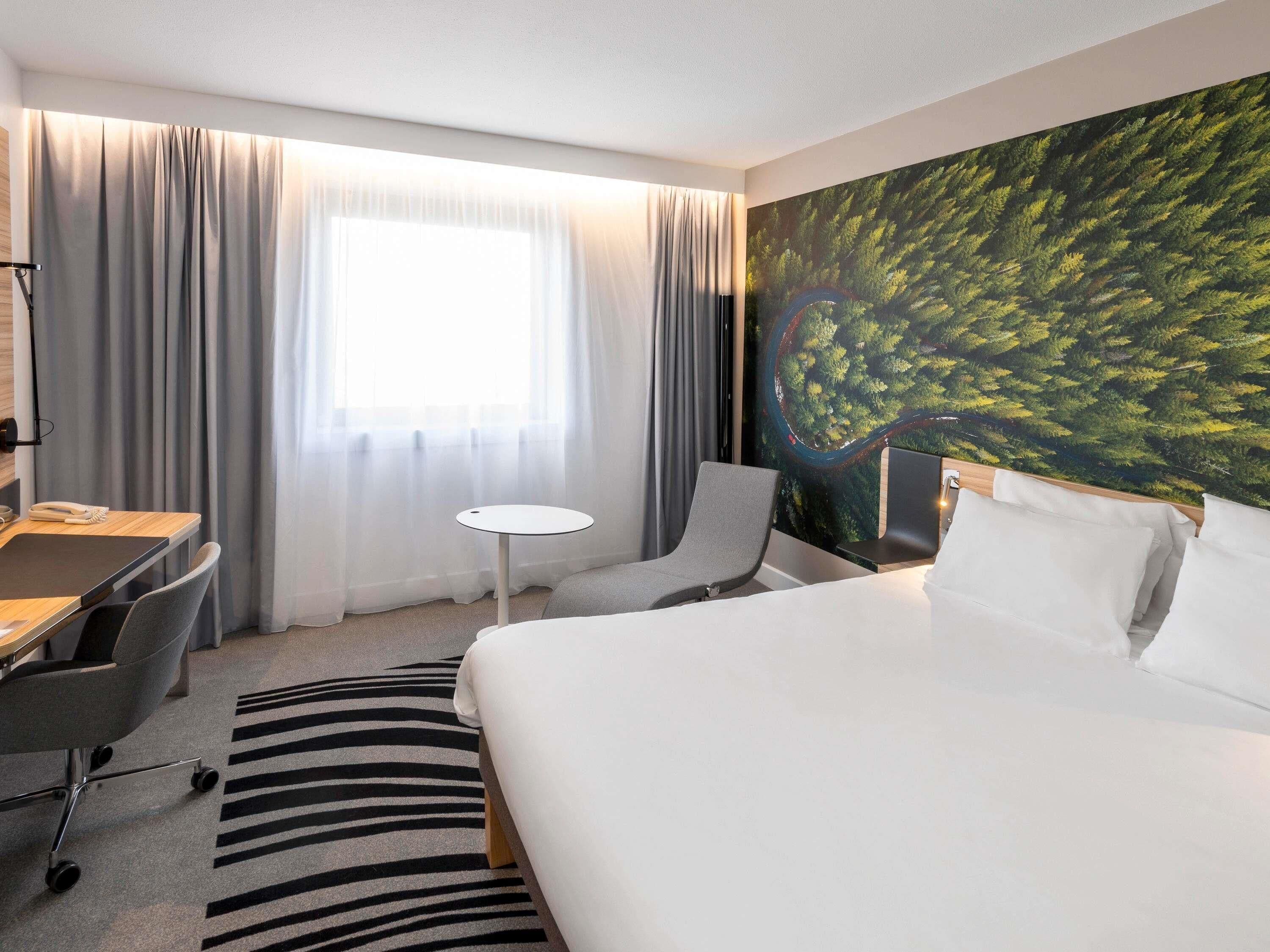 Hotellihuone, jossa on sänky, työpöytä, tuoli, pieni pöytä ja suuri ikkuna, jossa on verhot.