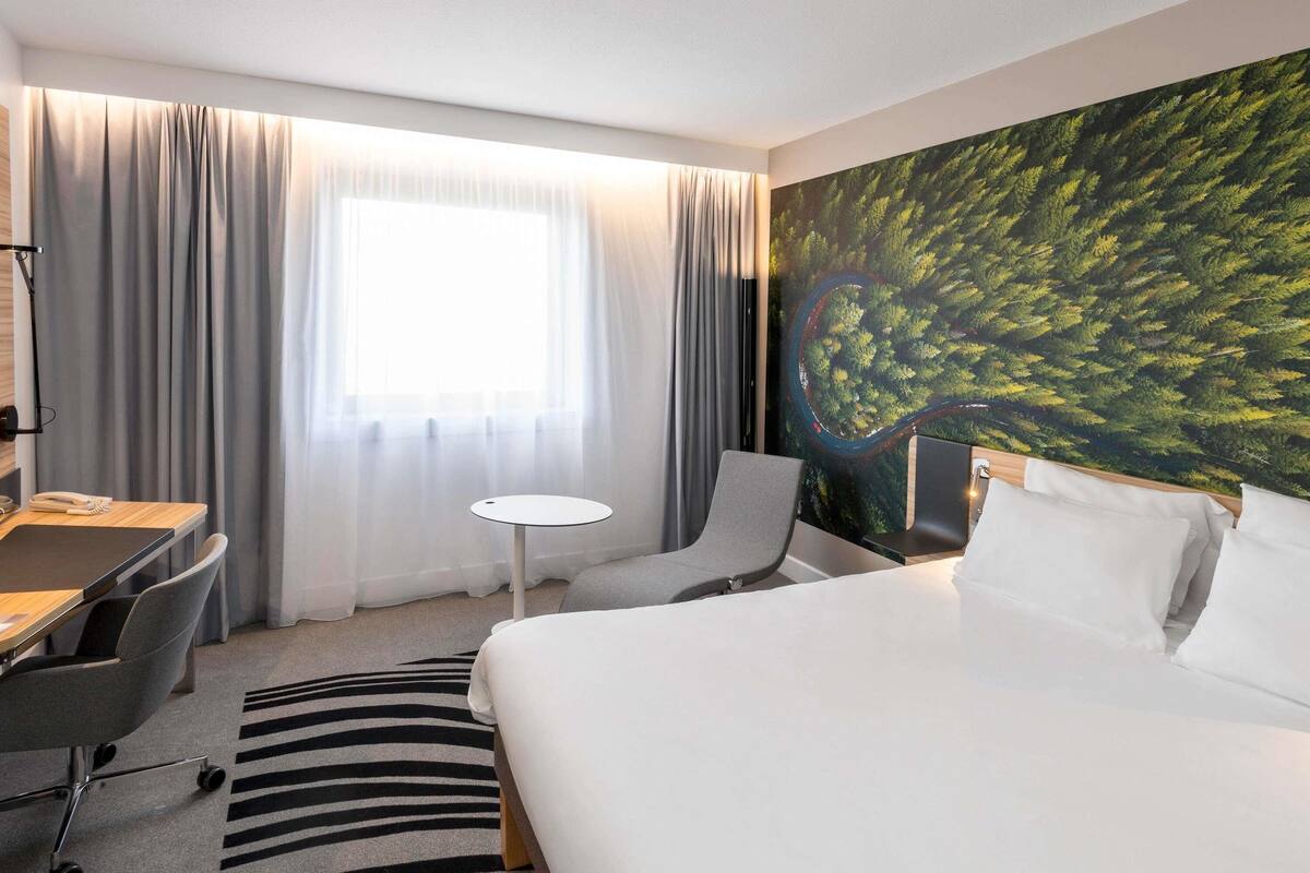 A hotel room with a bed, a desk, a chair, a small table, and a large window with curtains.