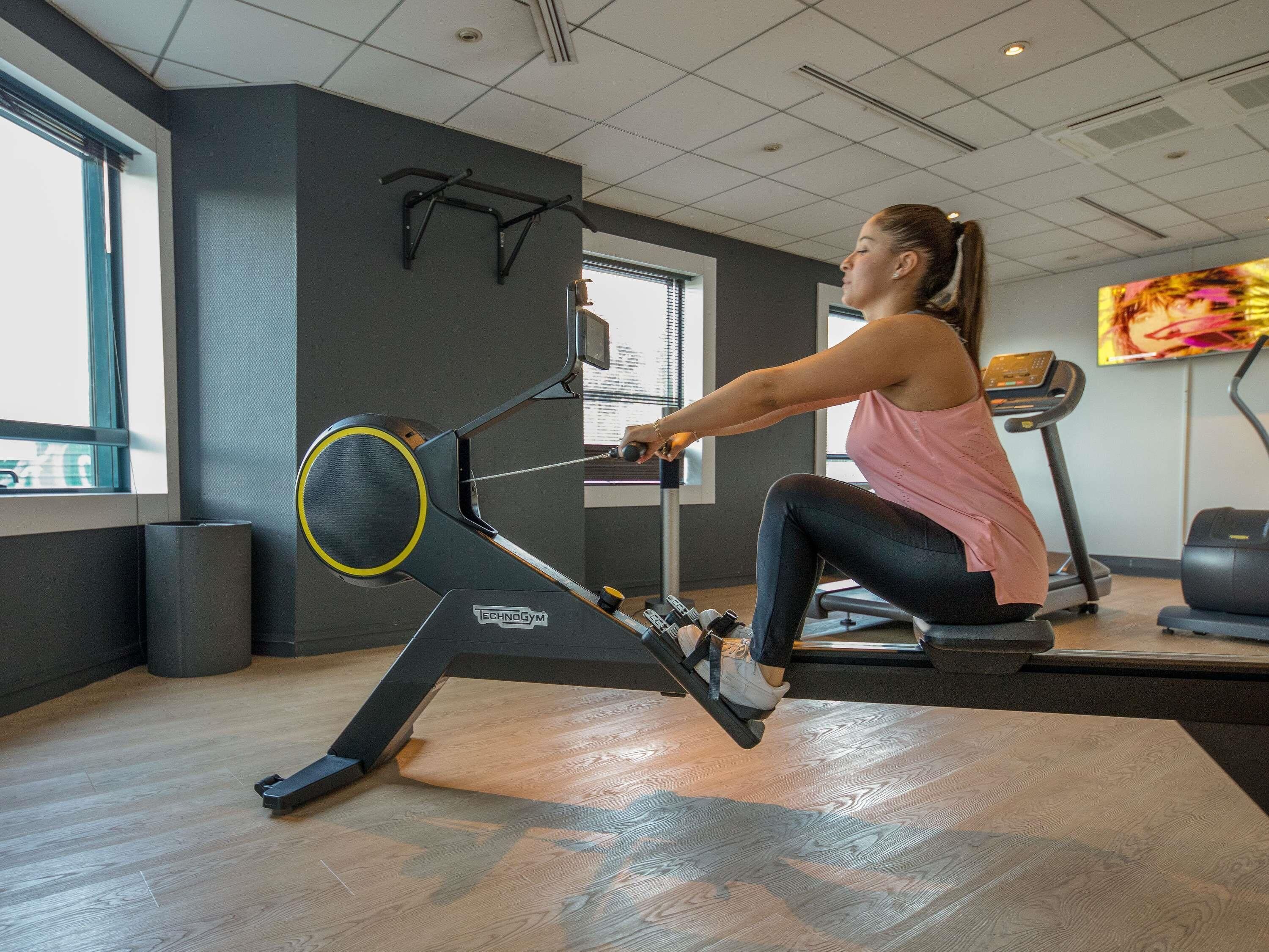 Une salle de sport équipée d’un rameur, d’un tapis de course et d’un écran de télévision diffusant une vidéo d’entraînement.