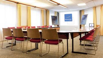 A conference room with a long table, red cushioned chairs, a projector screen displaying 'Novotel', and a banner with 'Welcome'.