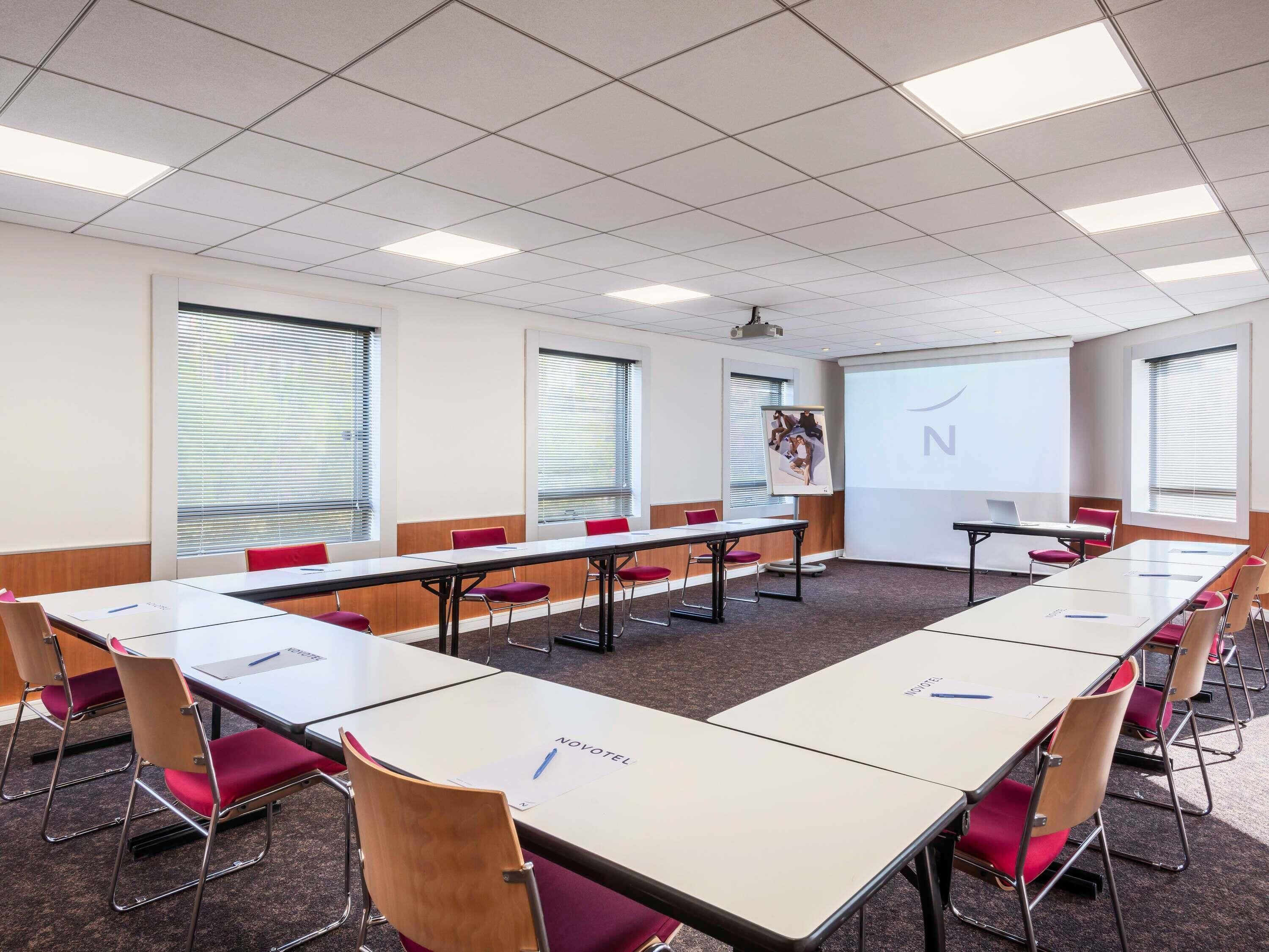 A conference room with multiple tables and chairs, a projector screen, and large windows with blinds.