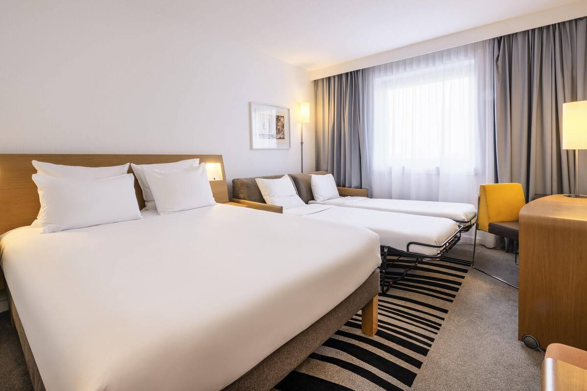A hotel room with two beds, a desk, a chair, a lamp, and a window with curtains.