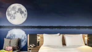 A hotel room with a bed, a nightstand, a chair, and a view of a lake and moon. - Novotel Marne-La-Vallée Noisy-Le-Grand (Noisy-le-Grand)
