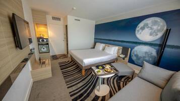 Quarto de hotel com cama, sofá, uma pequena mesa com comida, televisão e um quadro da lua.