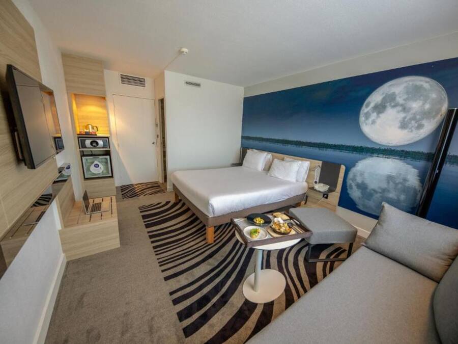 Quarto de hotel com cama, sofá, uma pequena mesa com comida, televisão e um quadro da lua.
