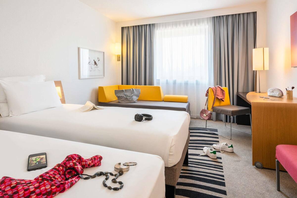 A hotel room with two beds, a sofa, a desk, and a chair. There is a window with curtains, a lamp, and a picture on the wall.