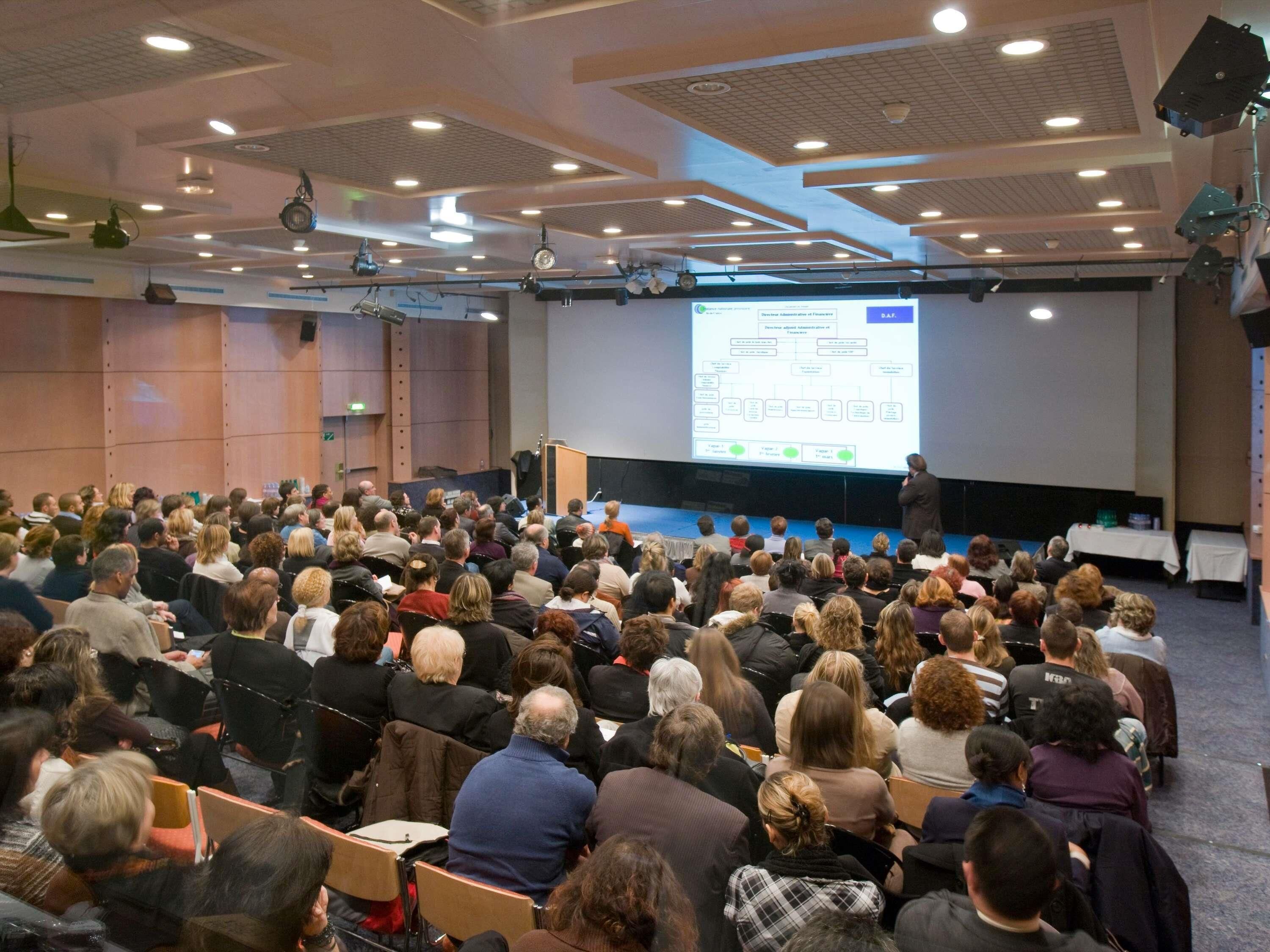 A conference room with a presentation screen, rows of chairs, and a stage with a podium.
