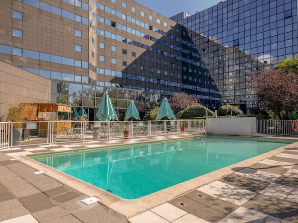 A swimming pool with a tiled deck, surrounded by a fence and umbrellas, with modern buildings in the background.
