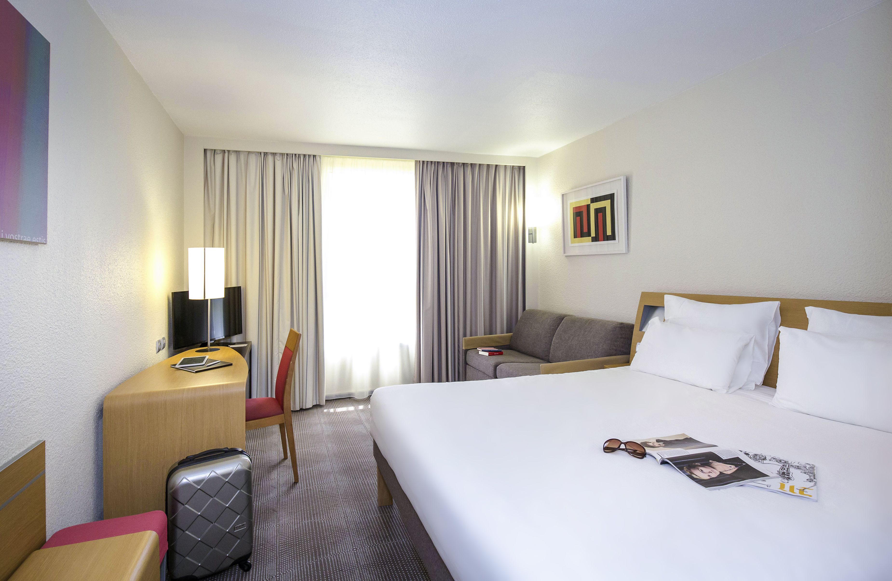 Habitación de hotel con cama, escritorio con lámpara, silla, televisor, maleta y ventana con cortinas.