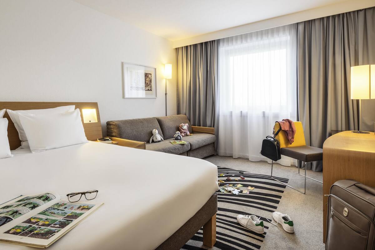 A hotel room with a bed, a sofa, a desk, a chair, a lamp, and a window with curtains.
