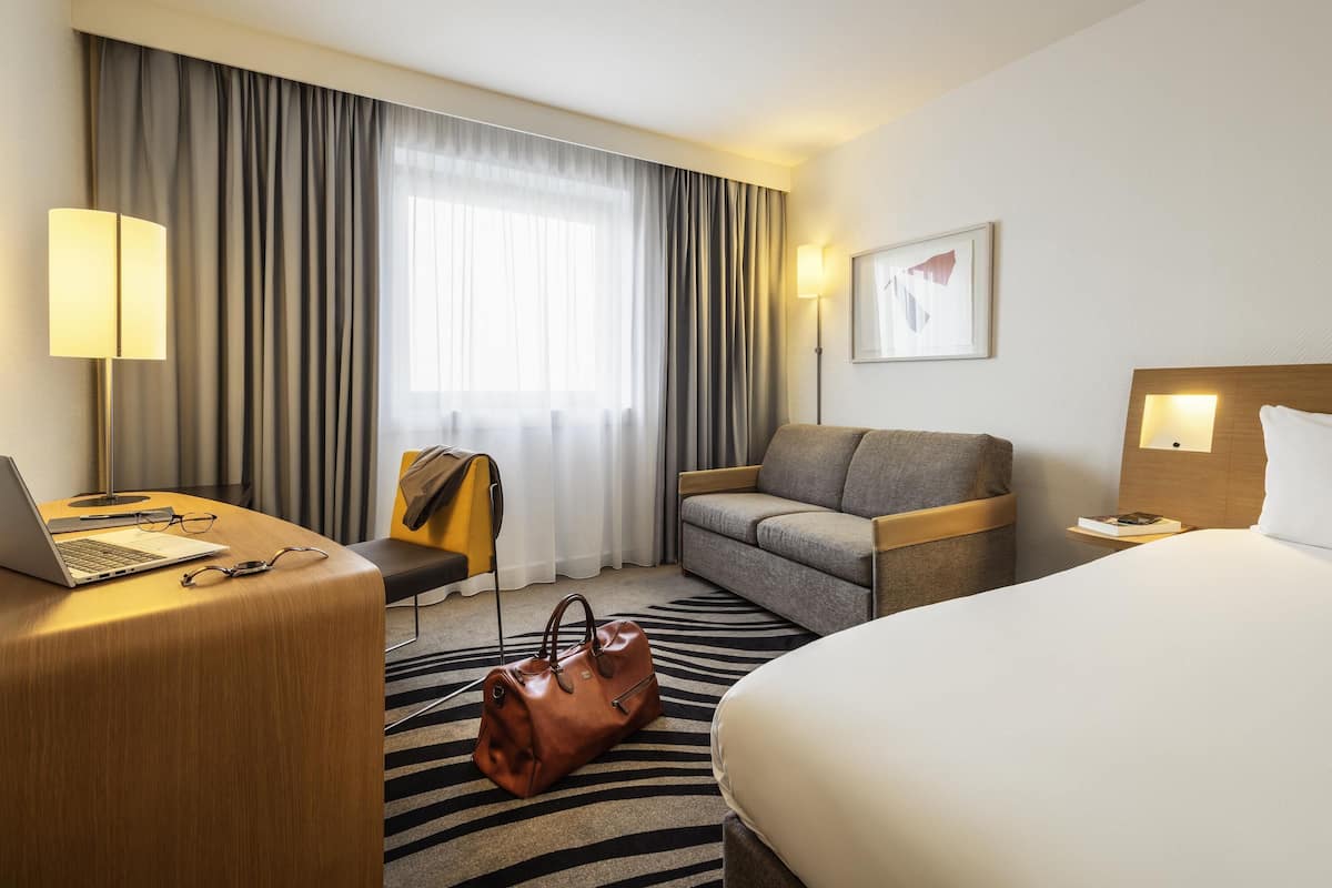 A hotel room with a bed, a desk with a laptop, a chair, a sofa, a nightstand, and a window with curtains.