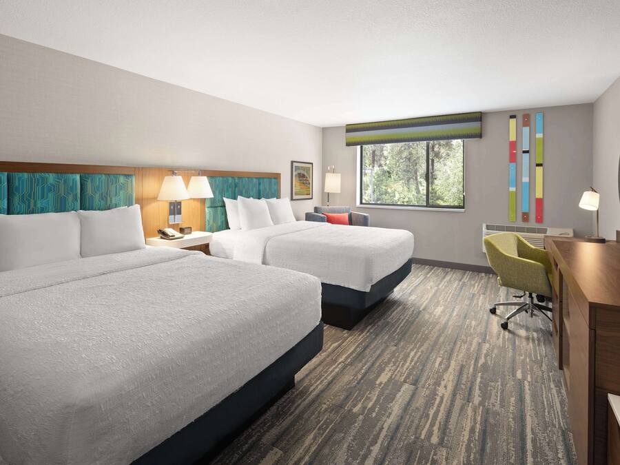A hotel room with two beds, a desk, a chair, a TV, and a window with a view of trees.