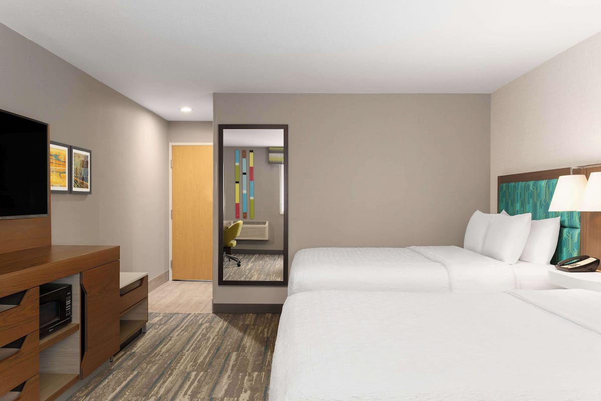 A hotel room with a bed, a desk with a TV, a mirror, and a door leading to a bathroom.