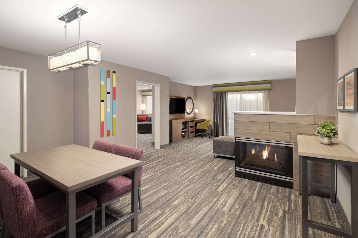 A modern hotel room with a dining area, a fireplace, a TV, and a desk.