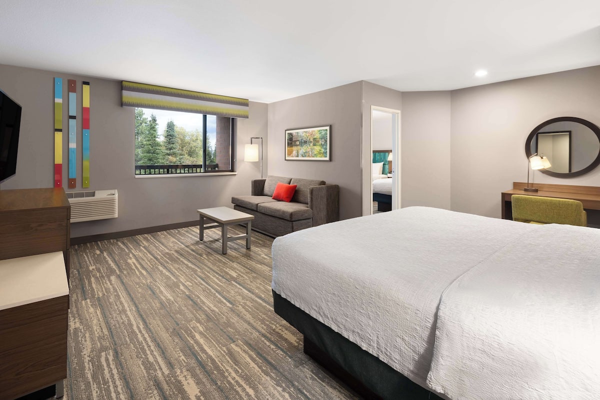 A hotel room with a large bed, a sofa, a small table, a TV, and a window with a view of trees.