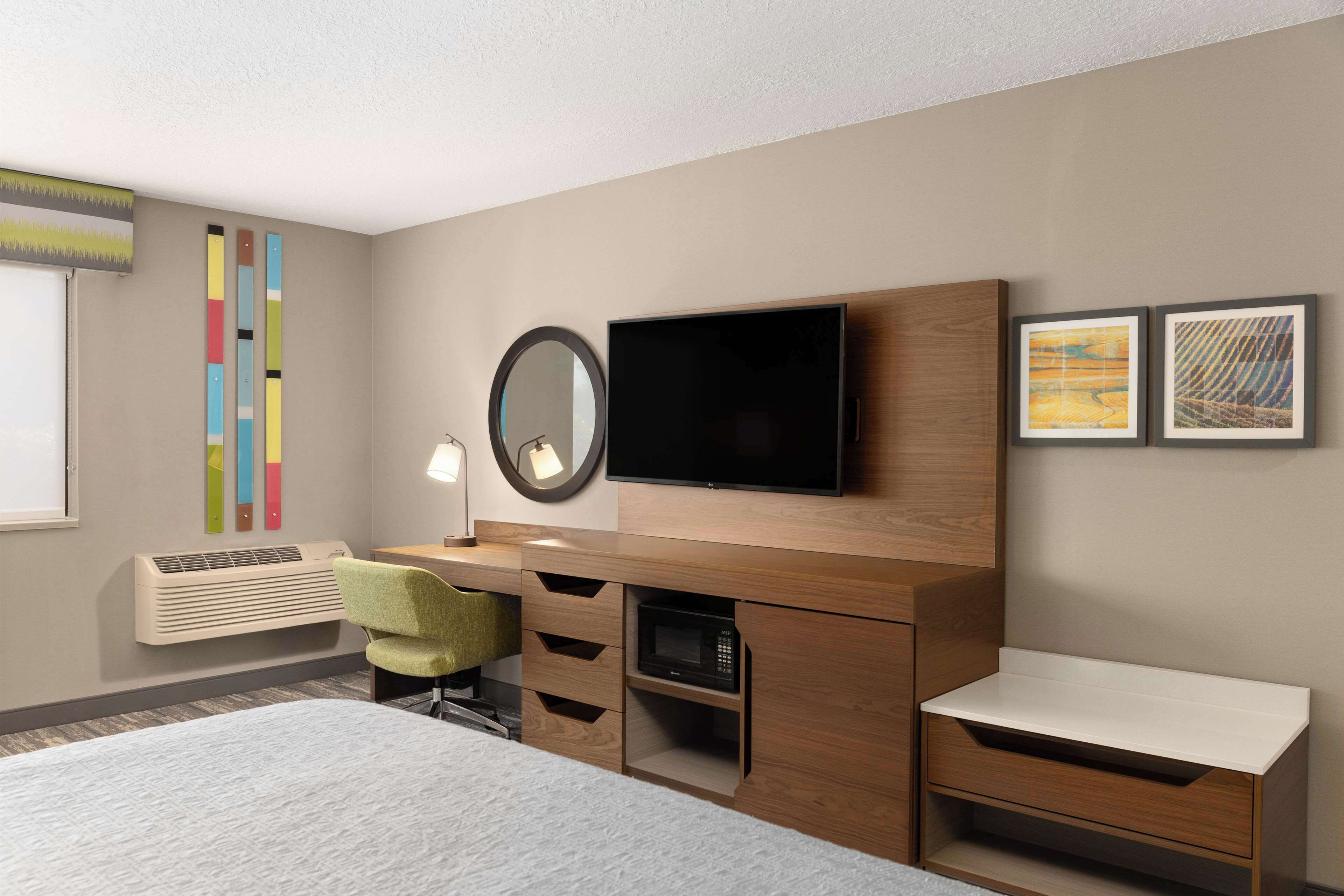 Una habitación de hotel moderna con una cama, un escritorio con silla, un televisor de pantalla plana, un espejo y obras de arte en la pared.