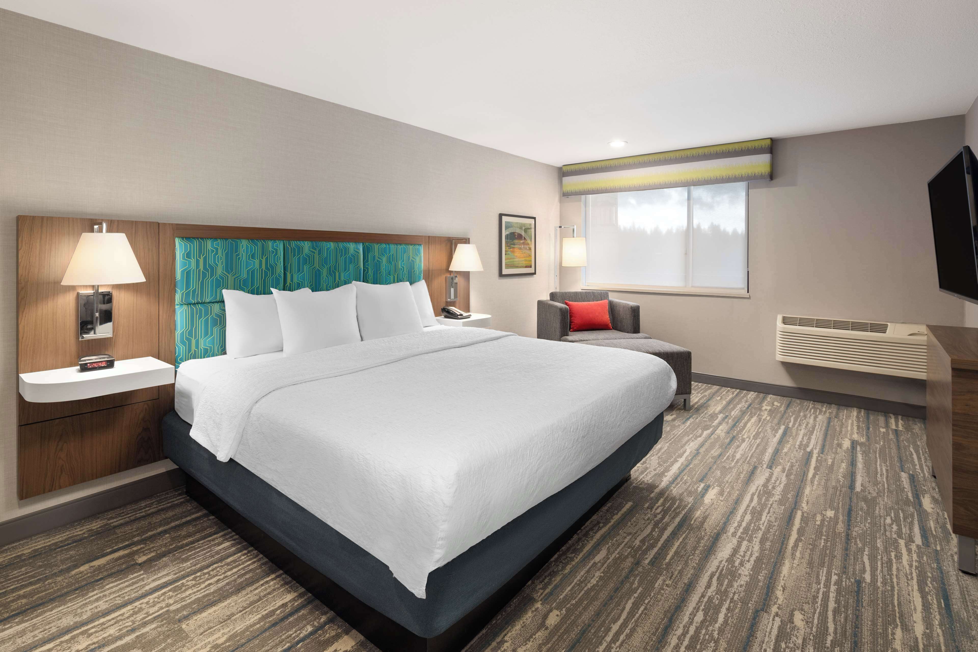 Quarto de hotel com uma cama grande, duas mesas de cabeceira, uma cadeira, uma televisão e uma janela.