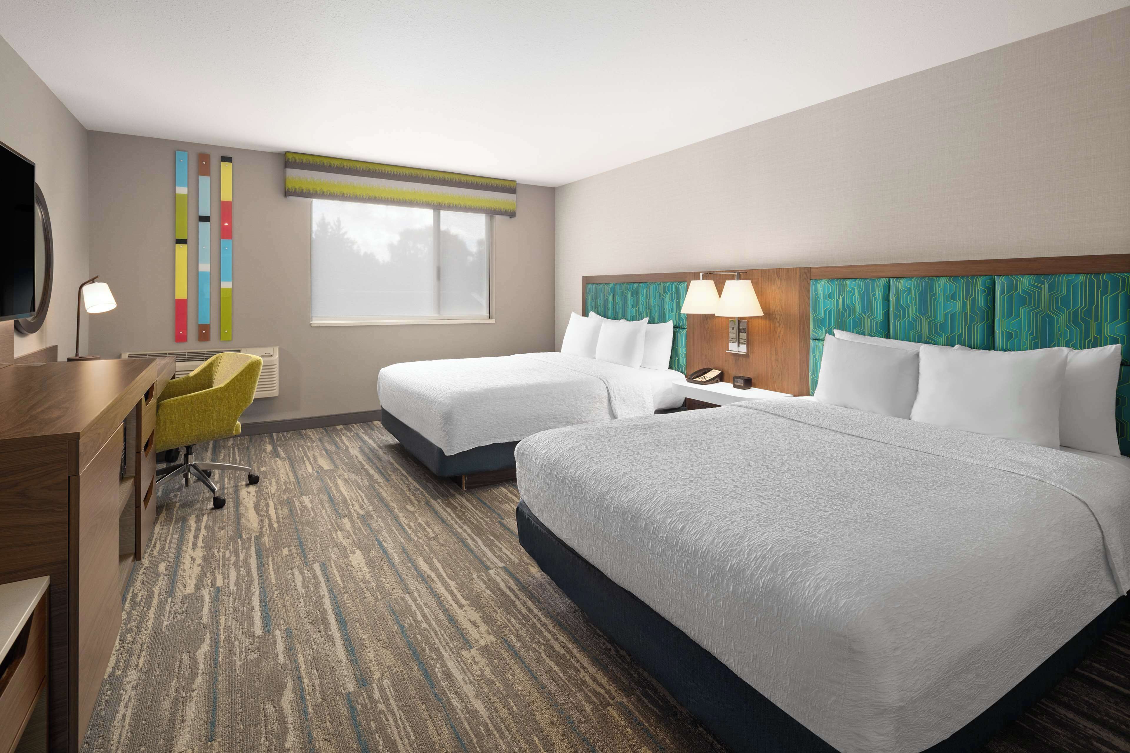 A hotel room with two beds, a desk, a chair, and a large window.