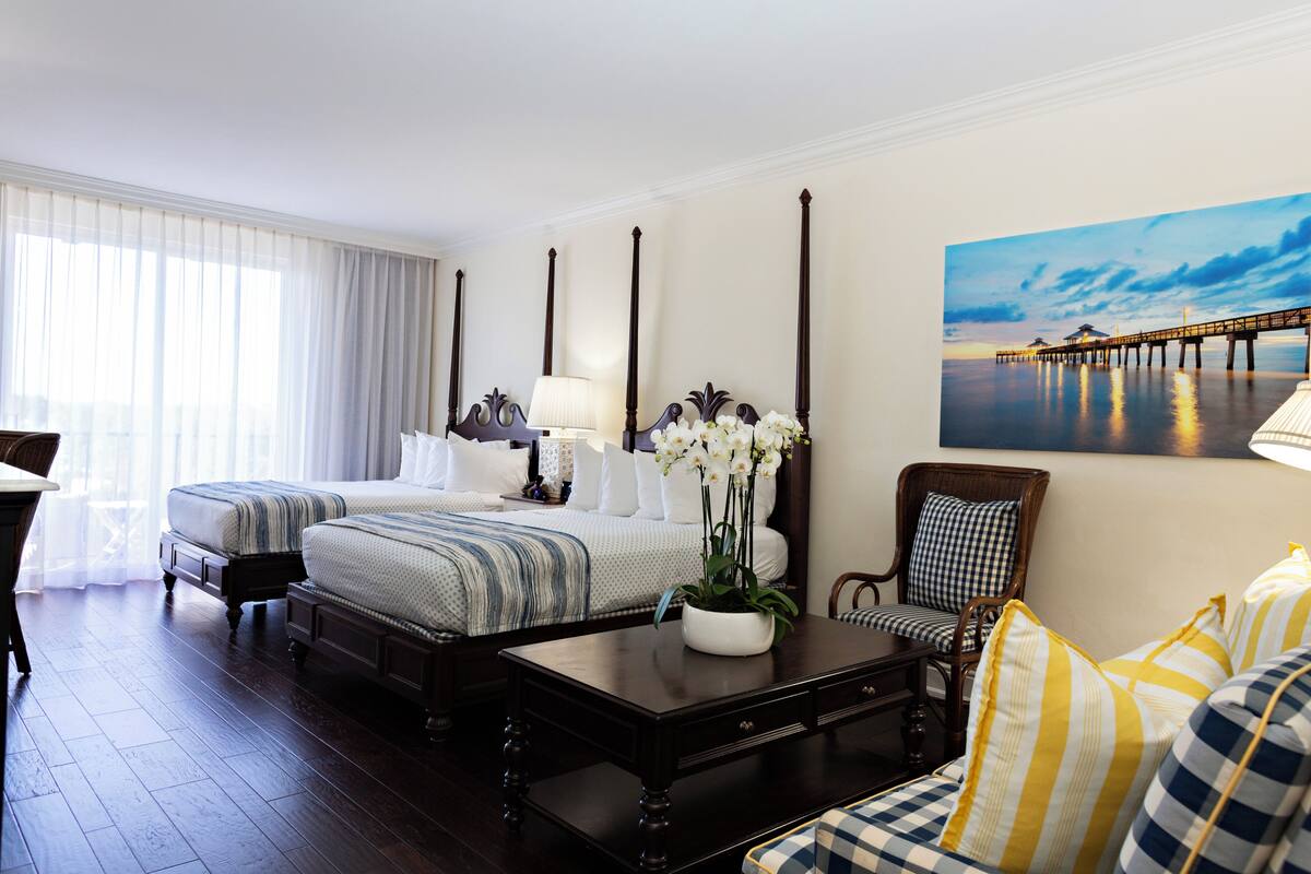 A hotel room with two beds, a sitting area with a sofa and armchairs, a coffee table, and a painting of a pier on the wall.