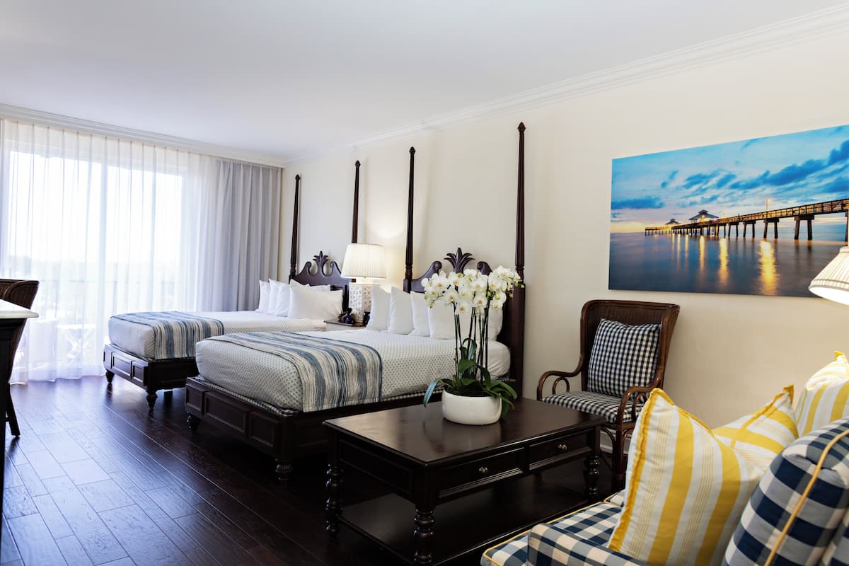 A hotel room with two beds, a sitting area with a sofa and armchairs, a coffee table, and a painting of a pier on the wall.