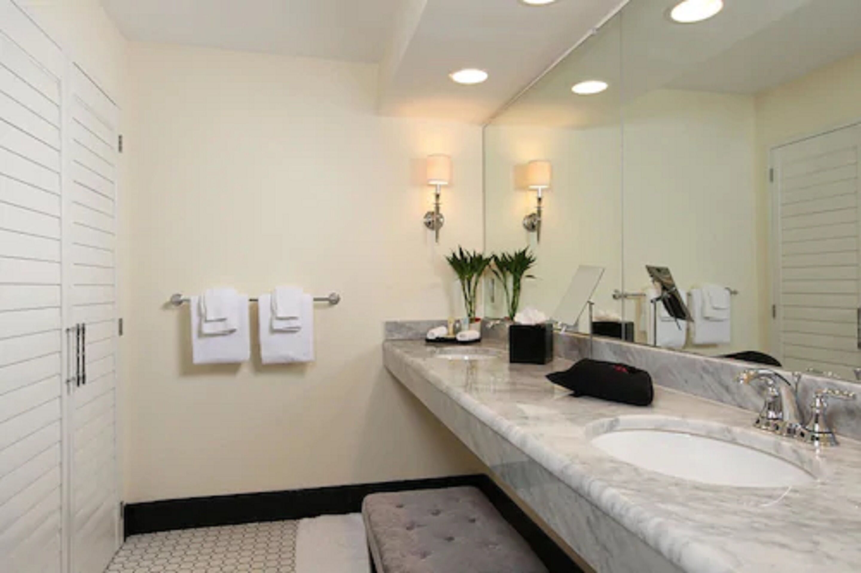 A bathroom with a large mirror, two towels hanging on the wall, a marble sink, and a small bench.