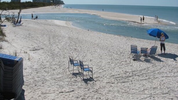 A beach with sand, a few chairs, and a person with an umbrella.
