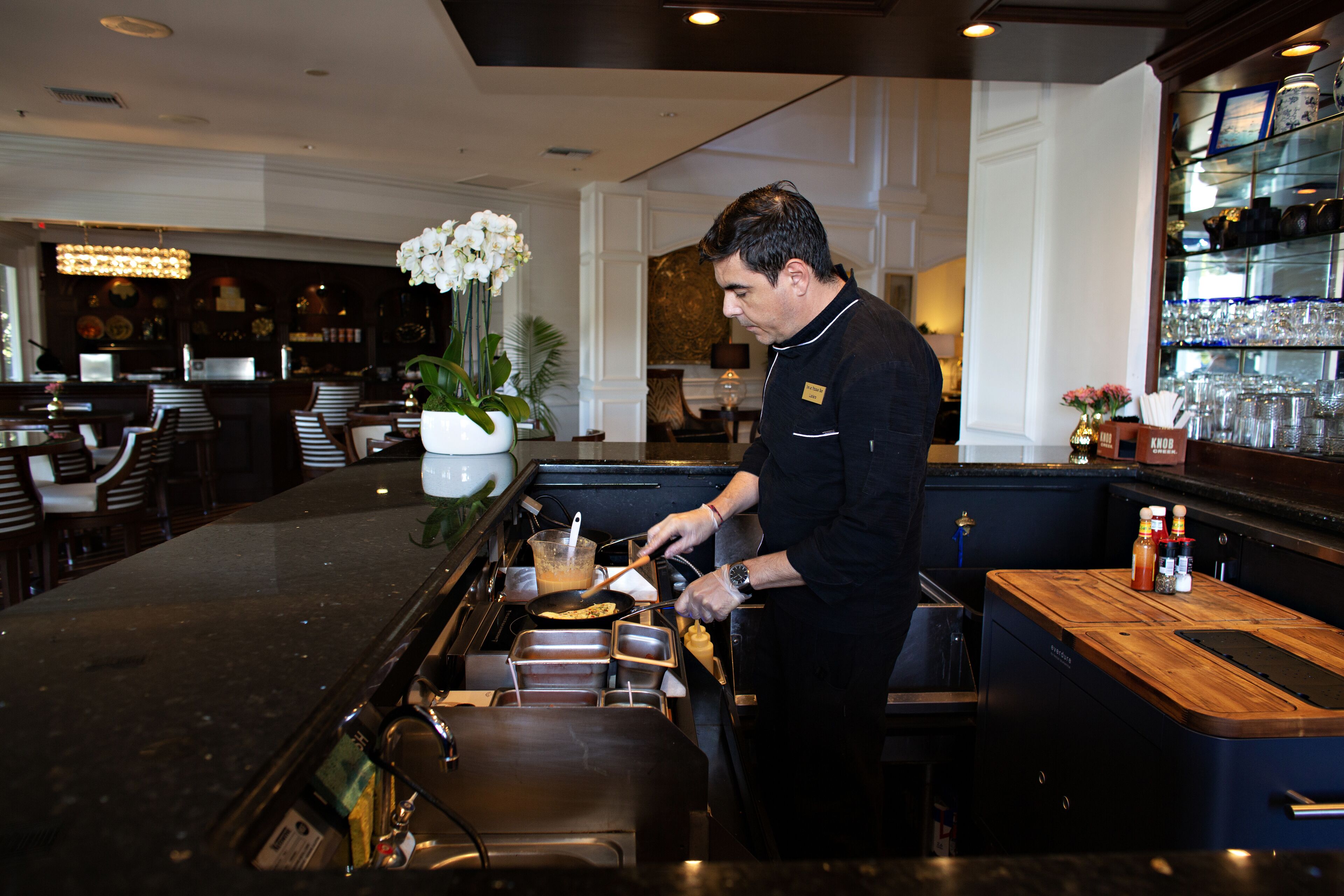 A bar area with a bartender preparing drinks, a potted plant, and a shelf with glassware.