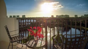 A balcony with a table and chairs, a vase with red flowers, and a view of a body of water and buildings in the distance.
