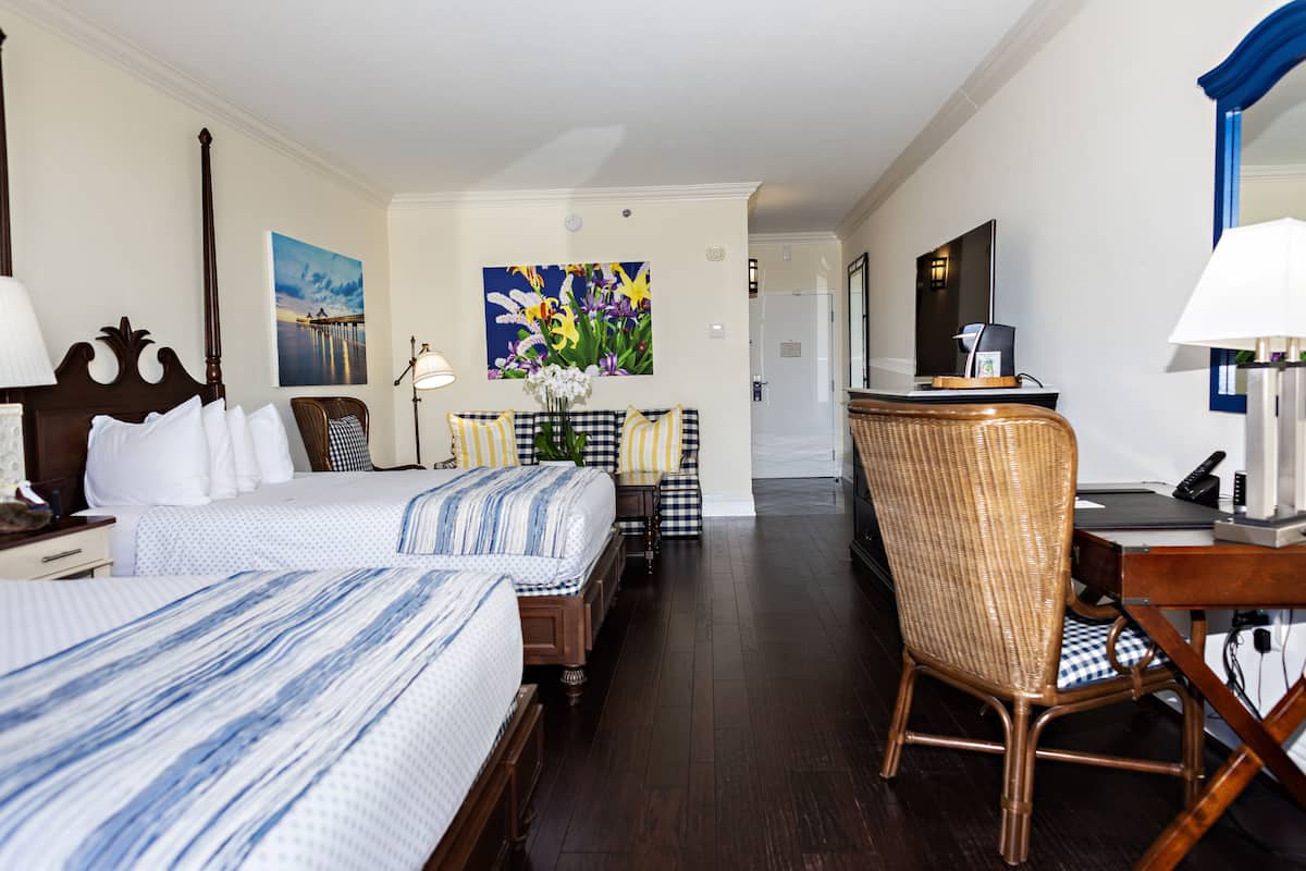 A hotel room with two beds, a desk, a chair, a table with a vase of flowers, and a painting on the wall.