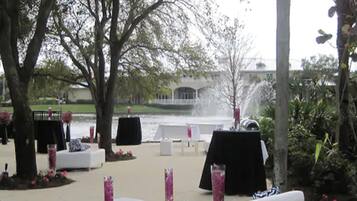 An outdoor seating area with white tables and chairs, surrounded by trees and a body of water.