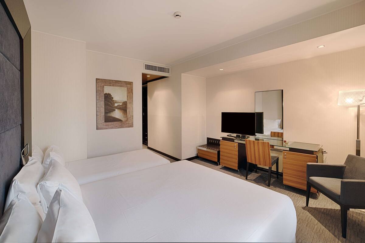 A hotel room with a large bed, a desk with a computer, a chair, a TV, and a painting on the wall.