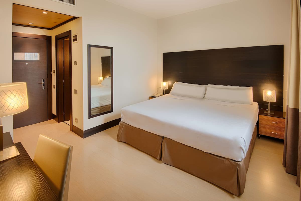 A hotel room with a large bed, a desk with a lamp, a mirror, and a door.