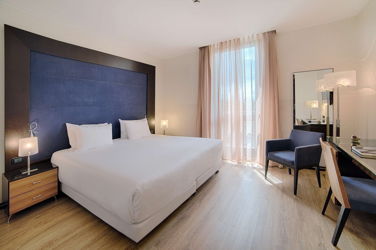A modern hotel room with a large bed, bedside lamps, a desk with a chair, and a large mirror.