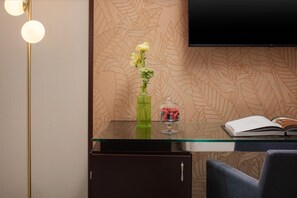 A hotel room with a glass desk, a vase with flowers, a fruit display, a book, and a flat-screen TV mounted on the wall.