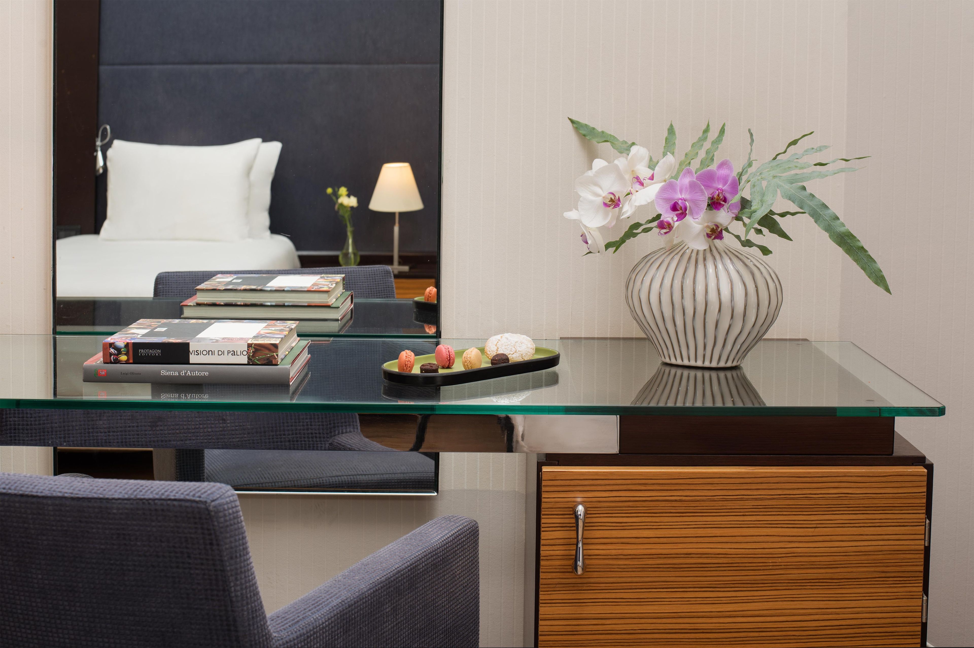A hotel room with a glass desk, a vase with flowers, a bed, and a nightstand with a lamp.
