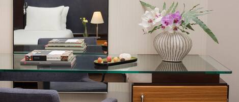 A hotel room with a glass desk, a vase with flowers, a bed, and a nightstand with a lamp.