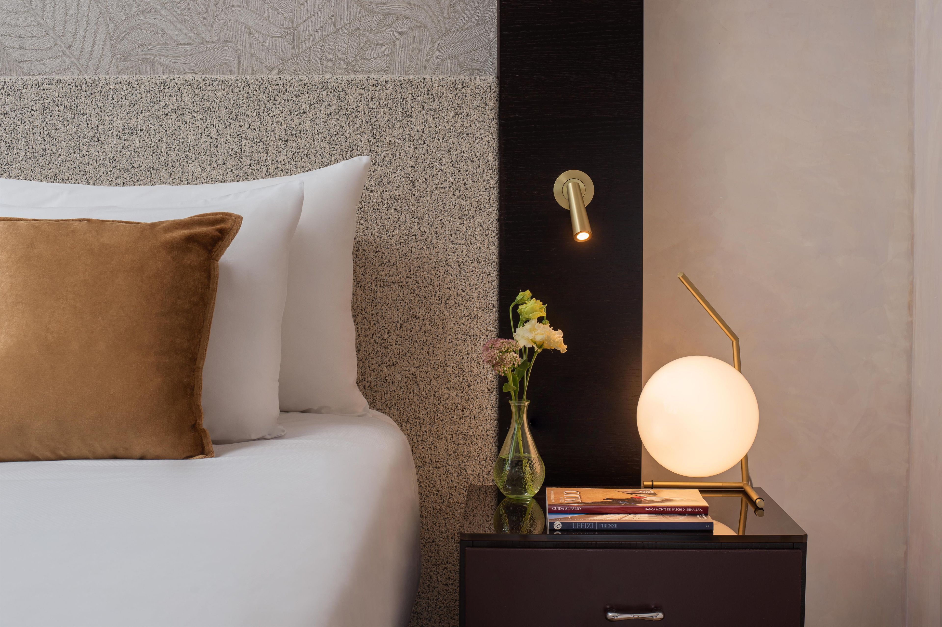 Una cama bien hecha con almohadas blancas y marrones, una mesita de noche con una lámpara, un florero con flores y un libro.