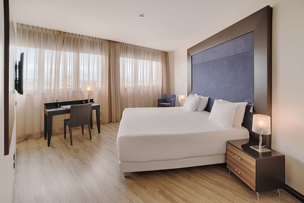 A hotel room with a large bed, a desk with a chair, a TV, and a bedside table with a lamp.