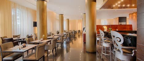 A spacious hotel lobby with multiple dining tables and chairs, large golden pillars, and a modern design.